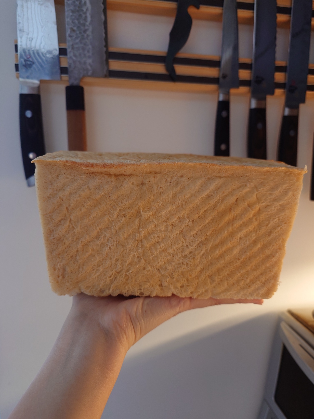 A rectangular loaf of bread being held up against a white wall with knife handles in the background