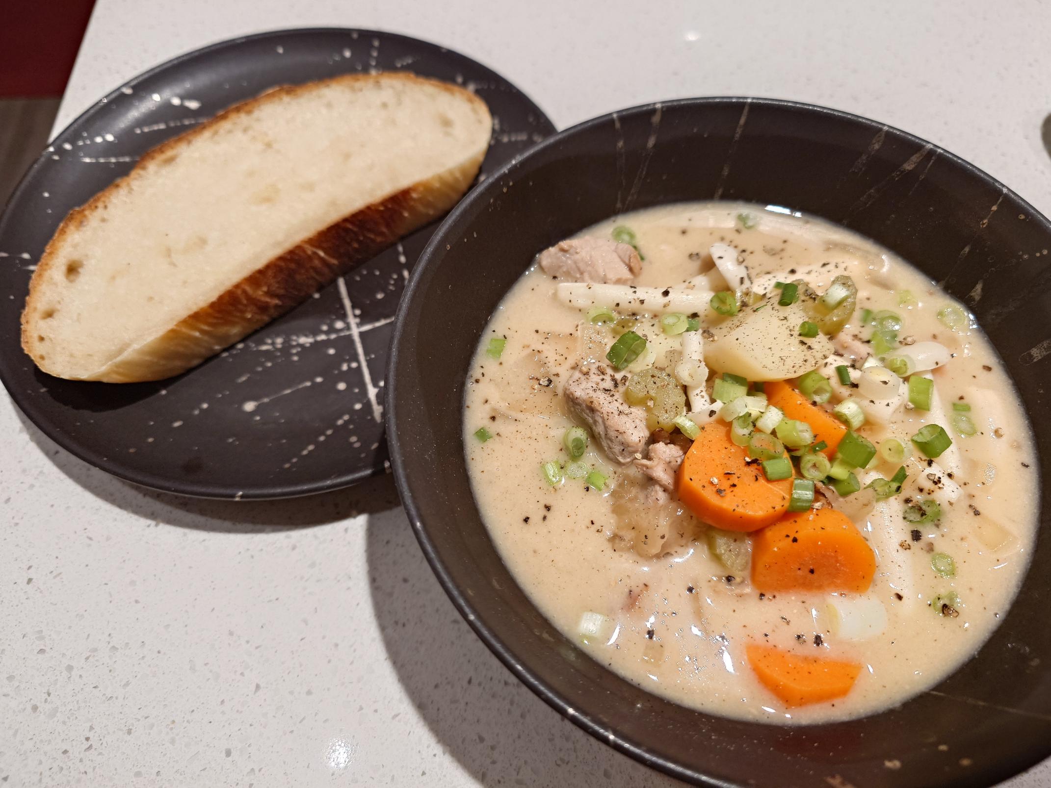 A creamy pork stew in a black bowl in the foreground. It has chunks of carrots visible. The background has a slice of bread on a black plate.